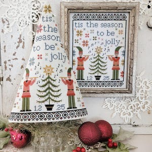 May include: A white cone-shaped Christmas tree ornament with a cross-stitch design featuring two figures playing musical instruments, a Christmas tree, and the text "tis the season to be jolly". The ornament is displayed on a glass stand with red ornaments and holly.