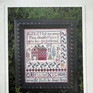 May include: A framed cross-stitch sampler titled "Jenny Bean's Christmas Sampler" by Shakespeare's Peddler. The sampler features a red house, trees, and text including "And had I but one penny...to buy gingerbread." The frame is dark brown.