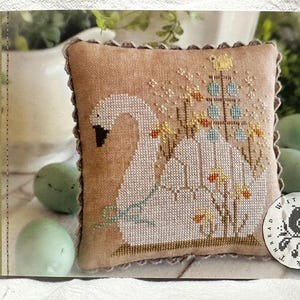 May include: A cross-stitch pattern featuring a white swan with a nest of eggs and a small tree with blue and yellow birds. The pattern is on a beige pillow with a scalloped edge. The text "Spring Swan" and "design by Brenda Gervais" is on the left side of the image. The text "#CS360" is on the right side of the image. The text "With Thread & Needle" is in the lower right corner of the image.