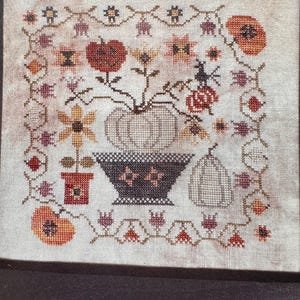 May include: Cross-stitch embroidery featuring a fall harvest theme. The design includes pumpkins, flowers, and a decorative bowl. The color palette consists of orange, brown, and beige, with intricate stitch details.