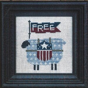 May include: A framed cross-stitch embroidery featuring a light blue sheep with white stars, wearing a shield with a white star and red and white vertical stripes. Above the sheep is a dark blue flag with the white text FREE in a block font.