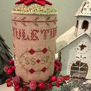 May include: A red and green Christmas-themed needlepoint pincushion with the words "Yuletide" and "Season's" stitched on it. The pincushion is decorated with a red bow and a red berry garland.