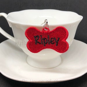 May include: Red bone-shaped dog tag with black embroidery that reads "Ripley".