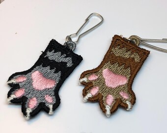 Op de afbeelding: Twee geborduurde pootvormige ritssluitingen met zilverkleurige metalen clips. De ene poot is zwart met grijze vachtdetails en roze kussentjes, de andere is bruin met beige vachtdetails en roze kussentjes. Beide hebben witte geborduurde klauwen.