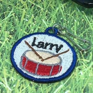 May include: A blue and white embroidered patch with a red drum and two drumsticks. The patch has the name "Larry" embroidered on it and a silver metal clip.