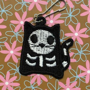 May include: Black and white embroidered cat skeleton zipper charm. The charm is attached to a silver zipper pull.