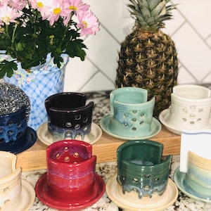 May include: A collection of eight ceramic dish soap holders in various colors, including blue, green, red, and white. The holders have a unique design with holes for drainage and a raised rim. They are displayed on a wooden surface with a pineapple in the background.