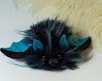 Pode incluir: Um morcego de pelúcia preto e azul-turquesa com olhos grandes e textura felpuda. O morcego tem um nariz pequeno e pontudo e orelhas grandes e arredondadas.