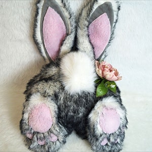 May include: A plush gray and white bunny rabbit with pink ears and paws. The bunny has a white fluffy tail and a pink flower on its back.
