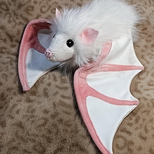 Op de afbeelding: Een witte pluche vleermuis met roze accenten op de oren en vleugelranden. De vleermuis heeft een pluizige kop en grote, zwarte ogen. De vleugels zijn wit met roze randen. Het speelgoed is ontworpen om op een vleermuis te lijken.