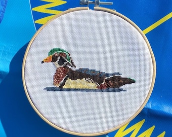 May include: A cross-stitch embroidery of a wood duck in a wooden hoop. The duck has a green head, brown body, and yellow and blue accents. The embroidery is set against a white background.