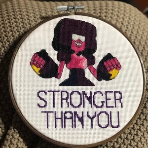 Puede incluir: Un aro de bordado de punto de cruz con una imagen de dibujos animados de Garnet del programa Steven Universe. Garnet lleva su atuendo característico y tiene los puños cerrados. El texto "Stronger Than You" está bordado debajo de la imagen.