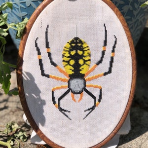 May include: A yellow and black spider with orange markings is embroidered on a white canvas in a wooden embroidery hoop.