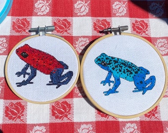 May include: Two embroidery hoops with cross-stitch designs of frogs. The frog in the left hoop is red with black spots and the frog in the right hoop is blue with black spots.