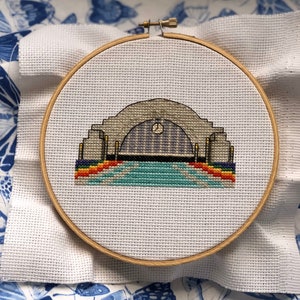 Könnte beinhalten: Ein fertiggestelltes Kreuzstich-Stickprojekt in einem Holzreifen, das ein stilisiertes Gebäude mit einem Regenbogenbogen darstellt.