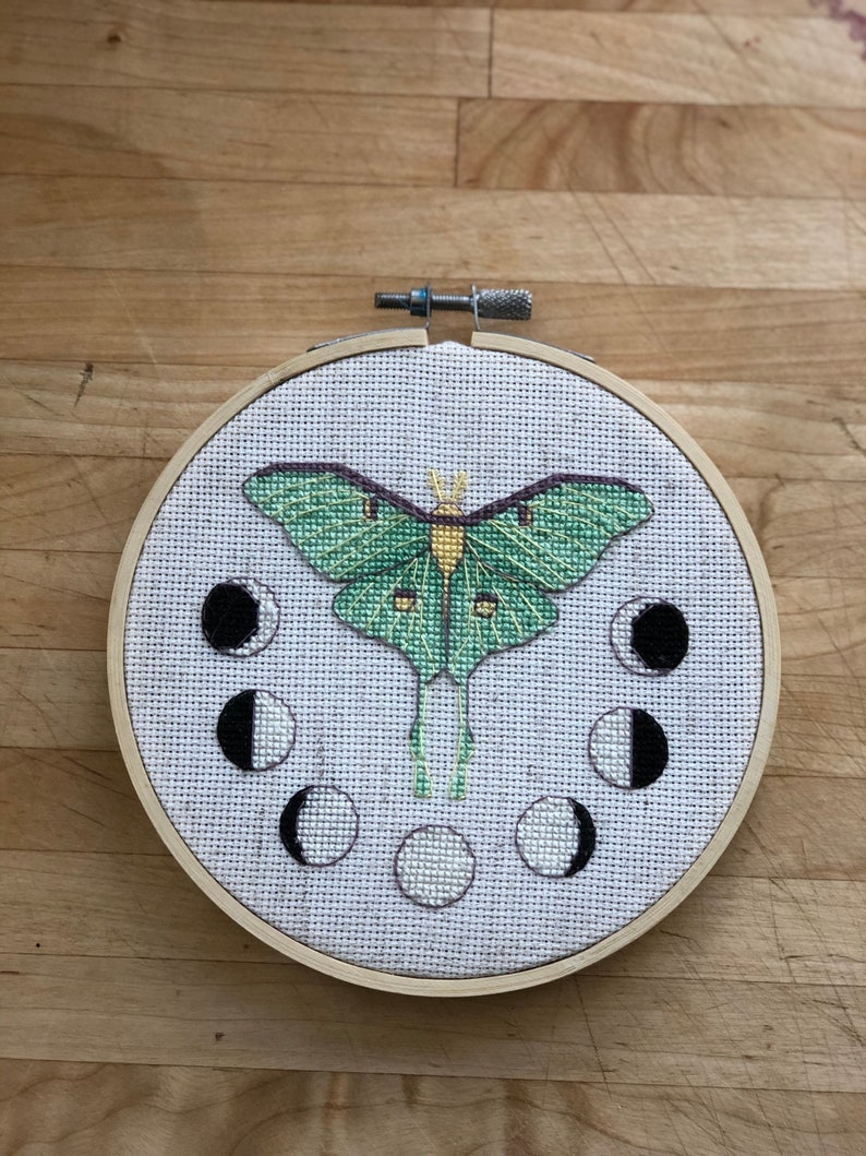 May include: A green and yellow luna moth embroidered on a white canvas with a wooden hoop. The moth is surrounded by eight black and white crescent moons.
