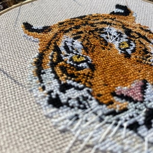 Puede incluir: Primer plano de un bordado de punto de cruz de la cara de un tigre. El tigre tiene rayas naranjas y negras, bigotes blancos y ojos amarillos. El bordado est&aacute; hecho en un lienzo blanco.