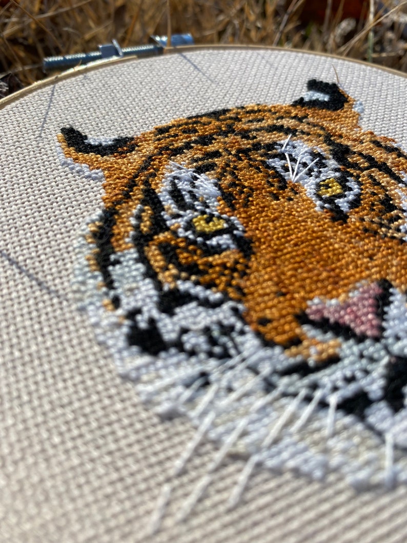 Puede incluir: Primer plano de un bordado de punto de cruz de la cara de un tigre. El tigre tiene rayas naranjas y negras, bigotes blancos y ojos amarillos. El bordado est&aacute; hecho en un lienzo blanco.