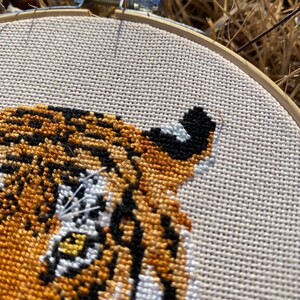 Puede incluir: Primer plano de un bordado de punto de cruz de la cara de un tigre. El ojo del tigre est&aacute; detallado con puntadas amarillas, blancas y negras. El bordado est&aacute; enmarcado en un aro de madera.