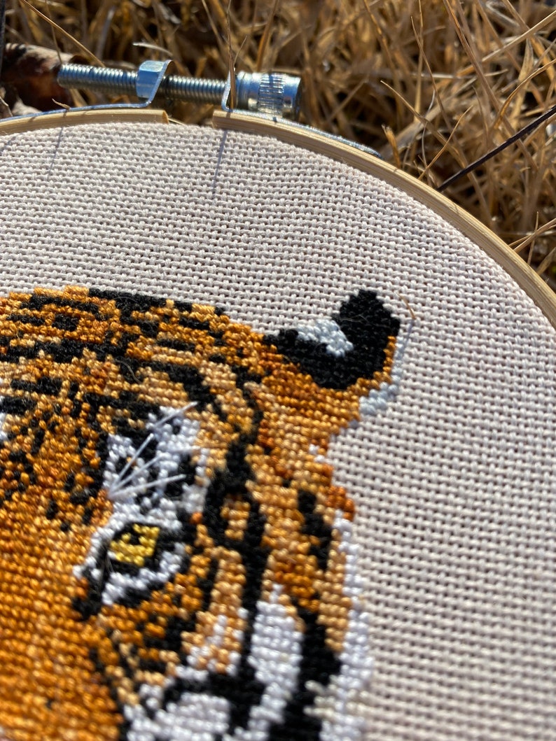 Puede incluir: Primer plano de un bordado de punto de cruz de la cara de un tigre. El ojo del tigre est&aacute; detallado con puntadas amarillas, blancas y negras. El bordado est&aacute; enmarcado en un aro de madera.