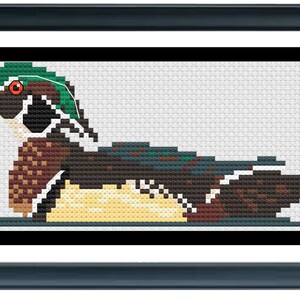 K&ouml;nnte beinhalten: Kreuzstich-Kunstwerk einer Wood Duck in einem rechteckigen Rahmen. Die Ente hat einen gr&uuml;nen Kopf, ein orangefarbenes Auge und einen mehrfarbigen K&ouml;rper. Der Rahmen ist dunkelblau und der Hintergrund ist wei&szlig;.