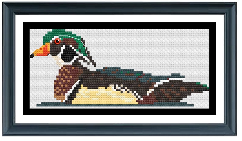 K&ouml;nnte beinhalten: Kreuzstich-Kunstwerk einer Wood Duck in einem rechteckigen Rahmen. Die Ente hat einen gr&uuml;nen Kopf, ein orangefarbenes Auge und einen mehrfarbigen K&ouml;rper. Der Rahmen ist dunkelblau und der Hintergrund ist wei&szlig;.