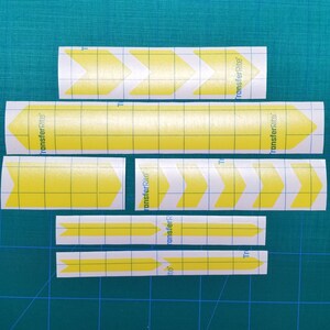 May include: A set of yellow arrow decals with "transferRite" printed on them. The decals are on a white gridded backing.