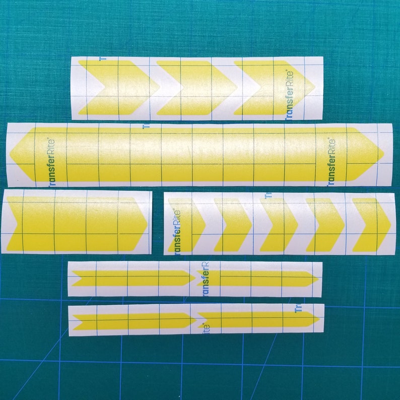 May include: A set of yellow arrow decals with "transferRite" printed on them. The decals are on a white gridded backing.