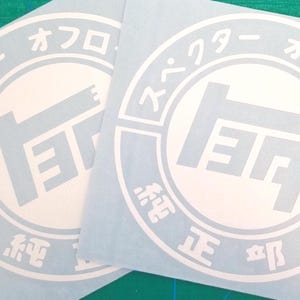 May include: Two square white vinyl decals featuring a circular emblem with a central stylized logo and Japanese text reading "スペクター オフロード" and "純正部品" arranged around the perimeter in a bold, clean font design.