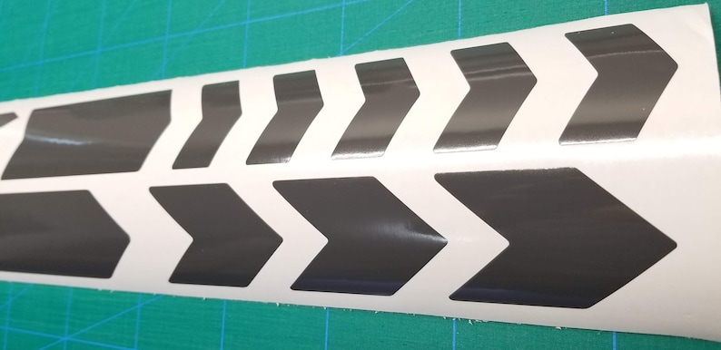 May include: A white vinyl sticker with a repeating pattern of black chevron arrows. The arrows are pointing to the right.