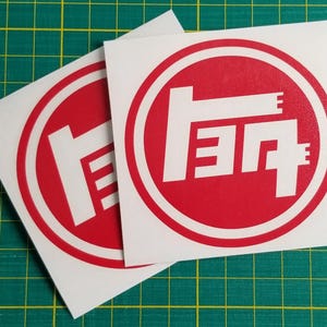 May include: Two square white vinyl decals featuring a red circular border and a stylized white Toyota logo in the center. The graphic design consists of bold geometric lines forming the Japanese characters for the automotive brand name.