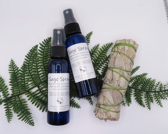 May include: Two blue glass bottles of sage spray with white labels and black text that reads "Sage Spray". The bottles are sitting on a white surface with a bundle of white sage smudge sticks and green fern leaves.