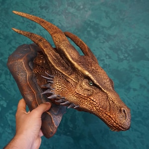 May include: A wall-mounted dragon head sculpture featuring a metallic bronze finish with intricate scale textures, long curved horns, and sharp spikes along the neck. The creature has a single visible green eye and a detailed snout.