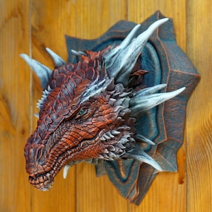 May include: A red and brown dragon head sculpture with silver horns and spikes, mounted on a dark blue and brown plaque. The dragon has green eyes and a textured, scaly skin.