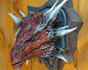 May include: A red and brown dragon head sculpture with silver horns and spikes, mounted on a dark blue and brown plaque. The dragon has green eyes and a textured, scaly skin.