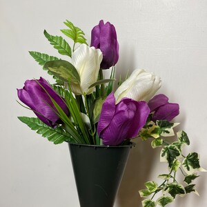 May include: A bouquet of purple and white tulips in a black cone vase. The vase is sitting on a brick surface. The tulips are artificial.