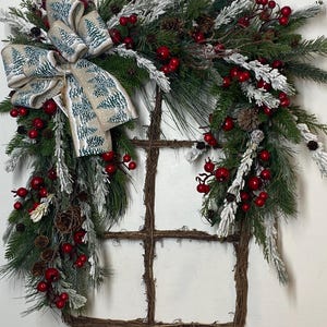 May include: A Christmas wreath with a rustic window frame base. The wreath features green pine branches, red berries, and frosted accents. A large bow with a winter tree pattern adds a decorative touch. Pinecones are also included.