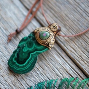 May include: A pendant necklace featuring a large, irregular slab of green malachite with concentric banding. The top is encased in ornate, coiled bronze-toned wirework holding a small, oval-shaped ruby zoisite cabochon on a brown cord.