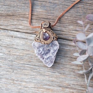 May include: A pendant necklace featuring a large, rough-cut, translucent purple amethyst crystal suspended from a tan leather cord. The top of the crystal is encased in intricate, coiled copper wire work that holds a smaller, polished amethyst stone.
