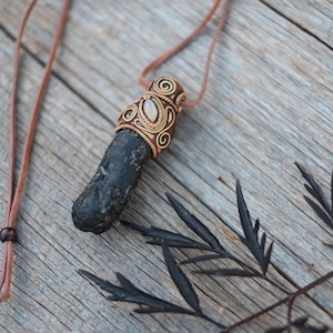 May include: A handcrafted necklace featuring a dark, elongated stone pendant. The pendant is adorned with intricate copper wirework and a small, oval-shaped stone. The necklace hangs from a brown leather cord, resting on a weathered wooden surface.