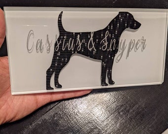 May include: A white rectangular tile with a black silhouette of a dog. The tile has the text "Cassius & Sniper" in a grey font.