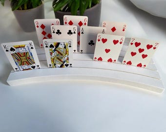 May include: White card holder with a curved design, holding a variety of playing cards, including a King of Spades, Queen of Hearts, 9 of Diamonds, 5 of Clubs, 6 of Hearts, 2 of Diamonds, Ace of Spades, 5 of Hearts, 7 of Hearts, and 7 of Spades.