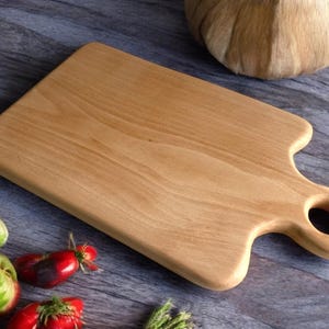May include: A light brown wooden cutting board with a rounded handle and a hole in the handle. The board is made of natural wood and has a smooth surface.