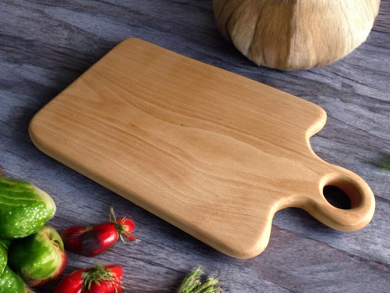 May include: A light brown wooden cutting board with a rounded handle and a hole in the handle. The board is made of natural wood and has a smooth surface.