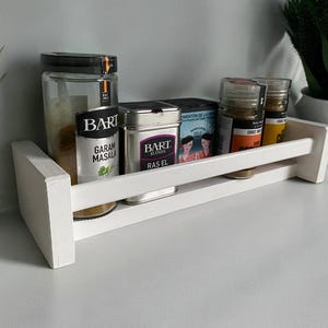 Puede incluir: Estante de especias de madera blanca con cinco tarros de especias. Los tarros contienen Garam Masala, Ras El Hanout y otras especias. El estante tiene un fondo blanco.