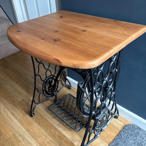 May include: A vintage sewing machine table with a wooden top and cast iron base. The tabletop is light brown and rectangular with rounded edges. The base is black, with decorative scrollwork and a foot pedal.