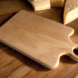 May include: A light brown wooden cutting board with a handle and a hole in the handle. The board is on a wooden surface.