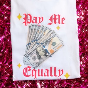 May include: White t-shirt with the text "Pay Me Equally" in red gothic font, accompanied by a fan of hundred-dollar bills. Yellow starburst graphics are placed around the text. The shirt is displayed on a pink, sparkly background.