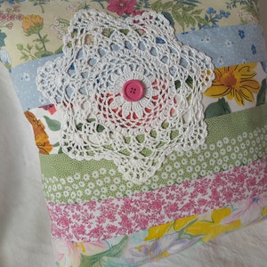 Peut inclure: Un coussin en patchwork avec une dentelle blanche au crochet au centre et un bouton rose. Le coussin est fait de différents tissus floraux dans des tons de rose, jaune, vert et bleu.