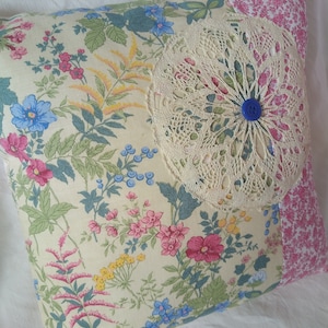 Peut inclure: Un oreiller de couleur crème avec un motif floral et une dentelle crochetée avec un bouton bleu au centre. L'oreiller est fait de chutes de tissu et a un motif floral rose et blanc au dos.